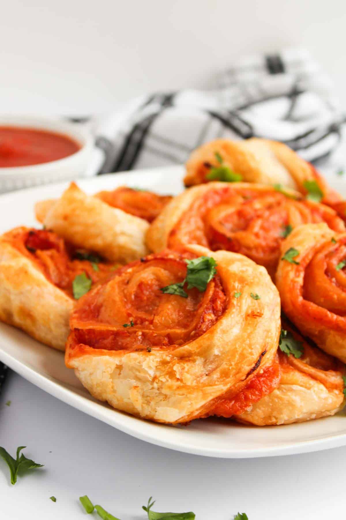 A plate of pizza pinwheels crafted from puff pastry, elegantly topped with fresh herbs. In the background, a bowl of tomato sauce awaits for dipping delight.