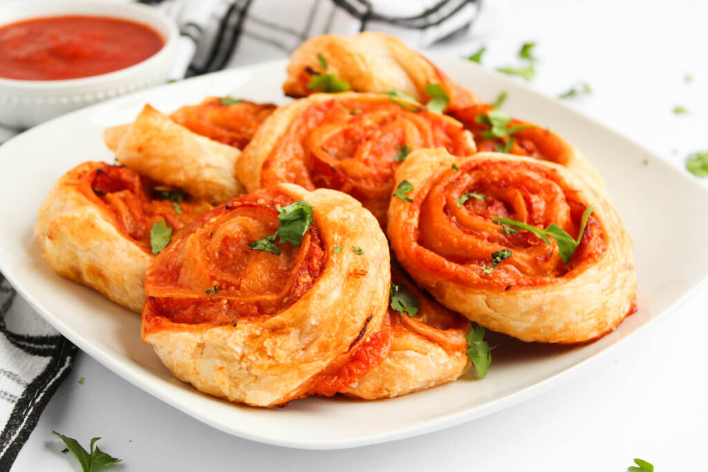 A white plate with several Pizza Pinwheels—puff pastry pinwheels filled with tomato sauce and cheese, garnished with parsley—sits next to a small bowl of marinara sauce.