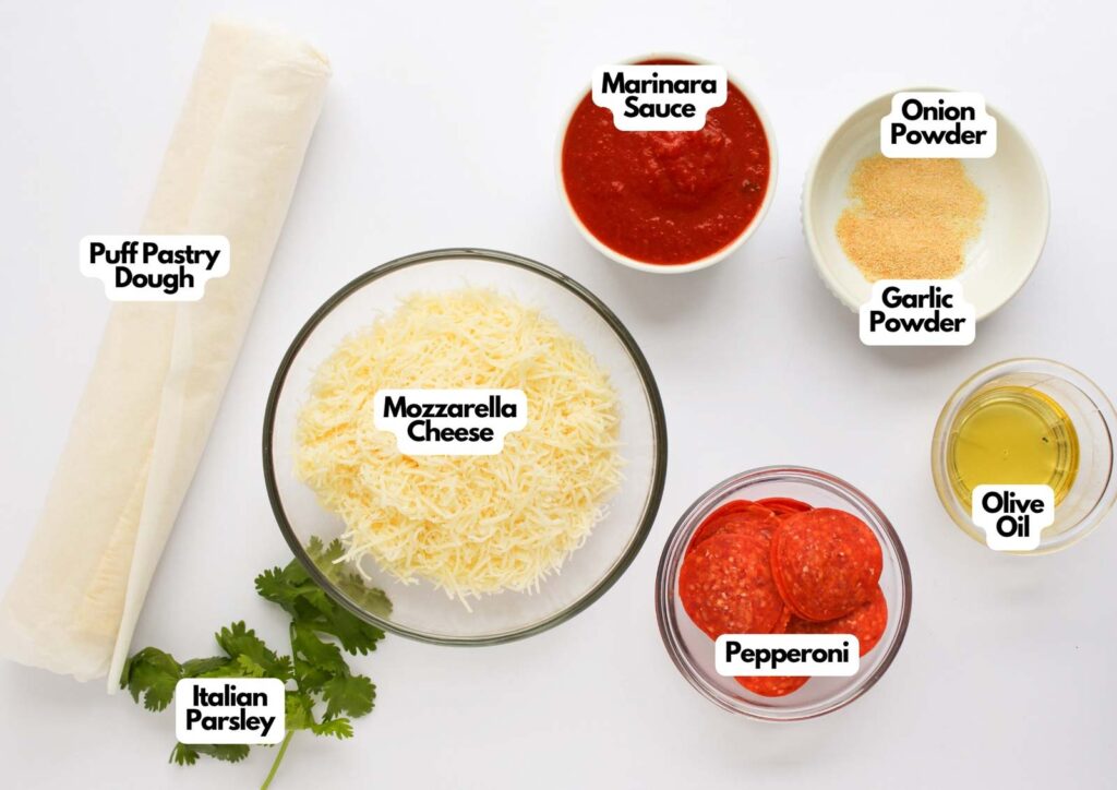 Ingredients for Pizza Pinwheels arranged on a white surface, featuring puff pastry dough, mozzarella cheese, marinara sauce, onion powder, garlic powder, olive oil, pepperoni, and Italian parsley.
