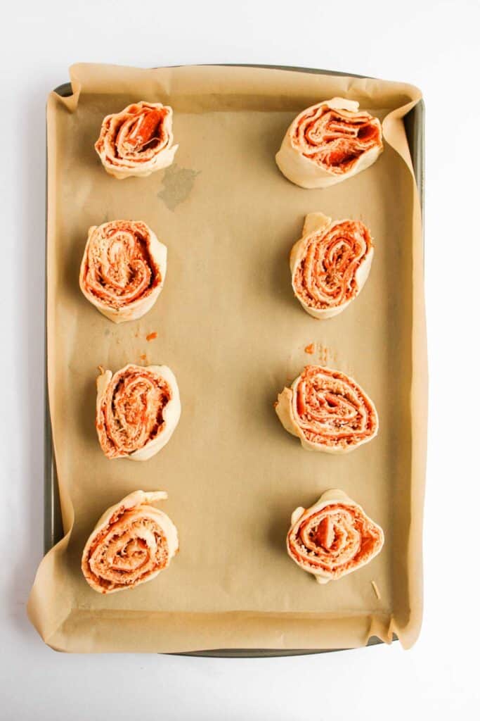 Eight uncooked Pizza Pinwheels with a tantalizing red filling are artfully arranged on a parchment-lined baking sheet, ready to bake.