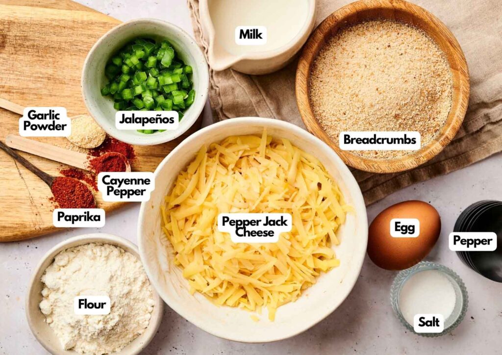 Ingredients for Air Fryer Rattlesnake Bites arranged on a surface: shredded cheese, jalapeños, breadcrumbs, egg, milk, flour, and spices like garlic powder, paprika, cayenne pepper, salt, and pepper.