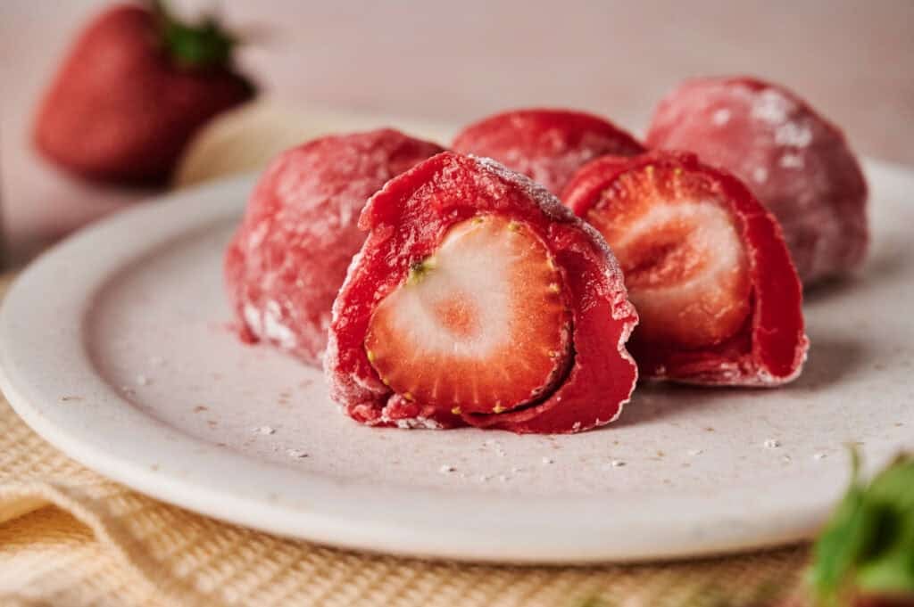 Close-up of Strawberry Mochi with one piece cut open, revealing a whole strawberry inside, all on a white plate.
