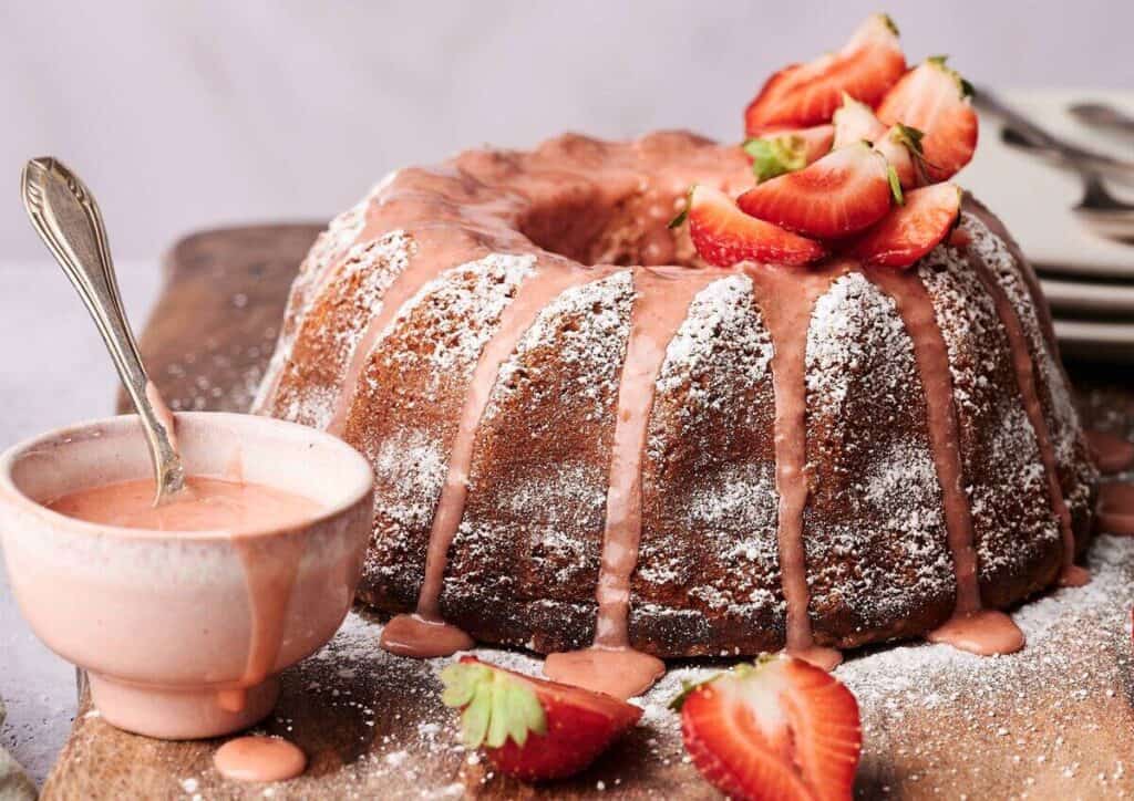 A bundt cake with pink icing and powdered sugar is topped with sliced strawberries, placed on a wooden board beside a bowl of pink glaze and more strawberries&mdash;perfect for pairing with your favorite ice cream cake.