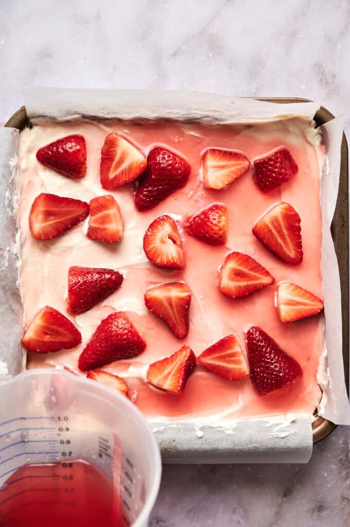 A baking pan lined with parchment holds a strawberry pretzel salad, its creamy layer topped with halved strawberries; a measuring cup with red liquid sits in the corner.