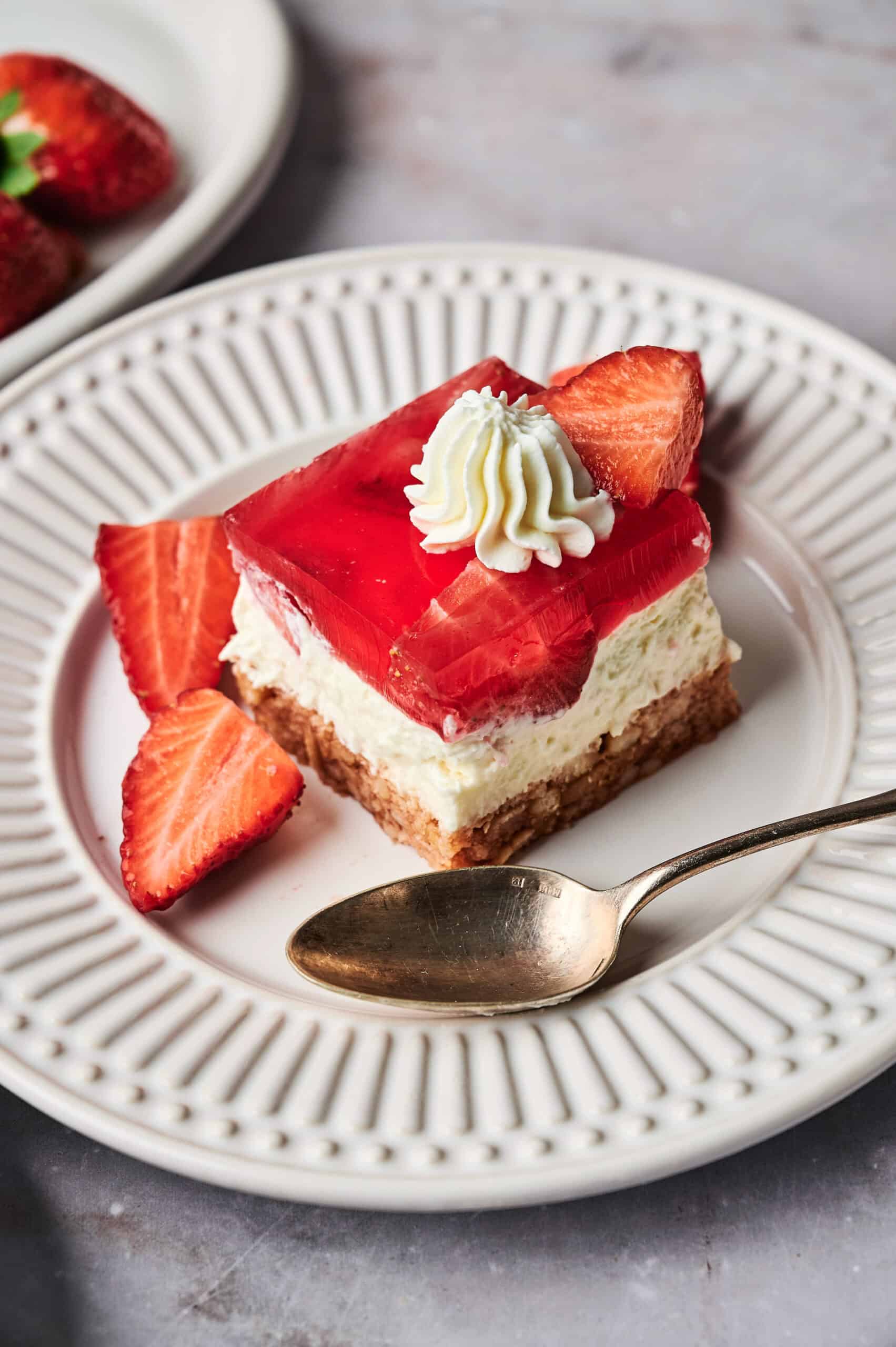 A slice of strawberry pretzel salad dessert with a crumb crust, creamy layer, red gelatin topping, and whipped cream, served on a white plate with a spoon and sliced strawberries.