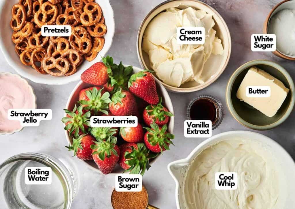 A top-down view of labeled ingredients for strawberry pretzel salad in bowls: pretzels, cream cheese, white sugar, butter, vanilla extract, Cool Whip, brown sugar, boiling water, strawberry Jello, and strawberries.