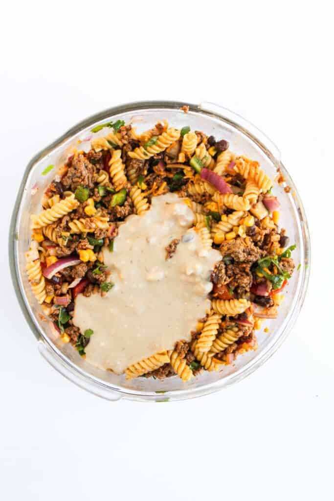 A glass dish filled with taco pasta salad featuring rotini, ground meat, vegetables, and topped with a creamy white sauce against a white background.