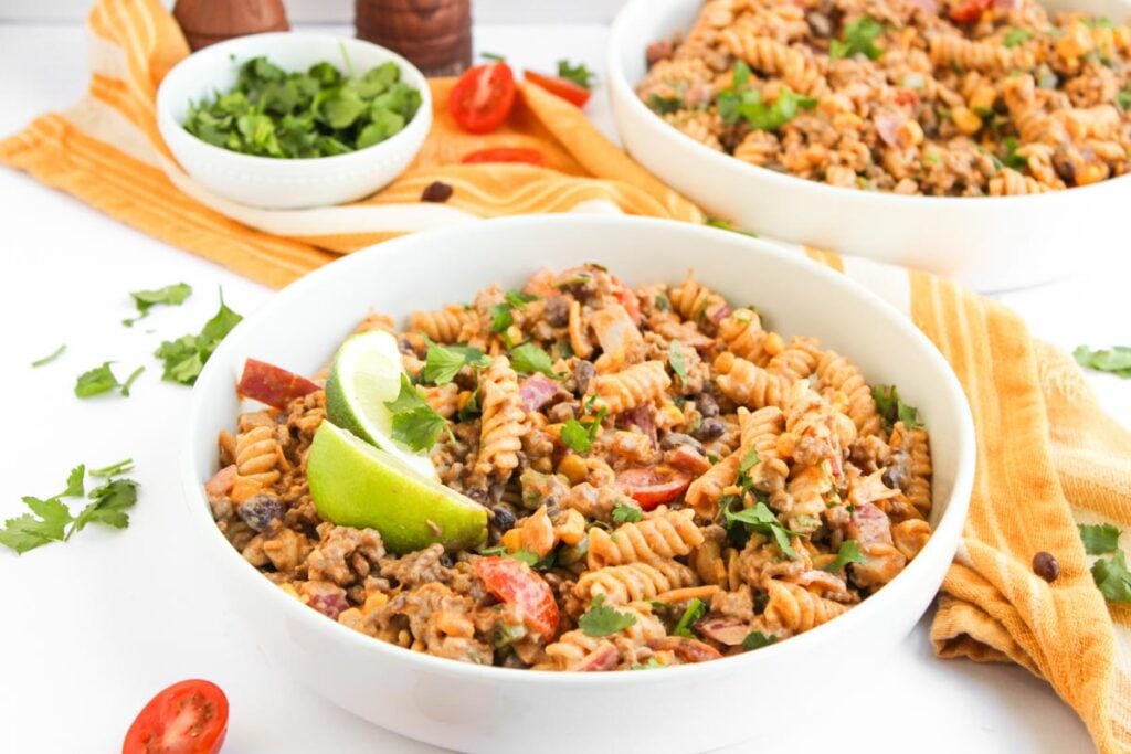 A bowl of creamy Taco Pasta Salad with rotini, vegetables, black beans, and cilantro, garnished with lime wedges. A second bowl and small dishes of herbs are in the background.