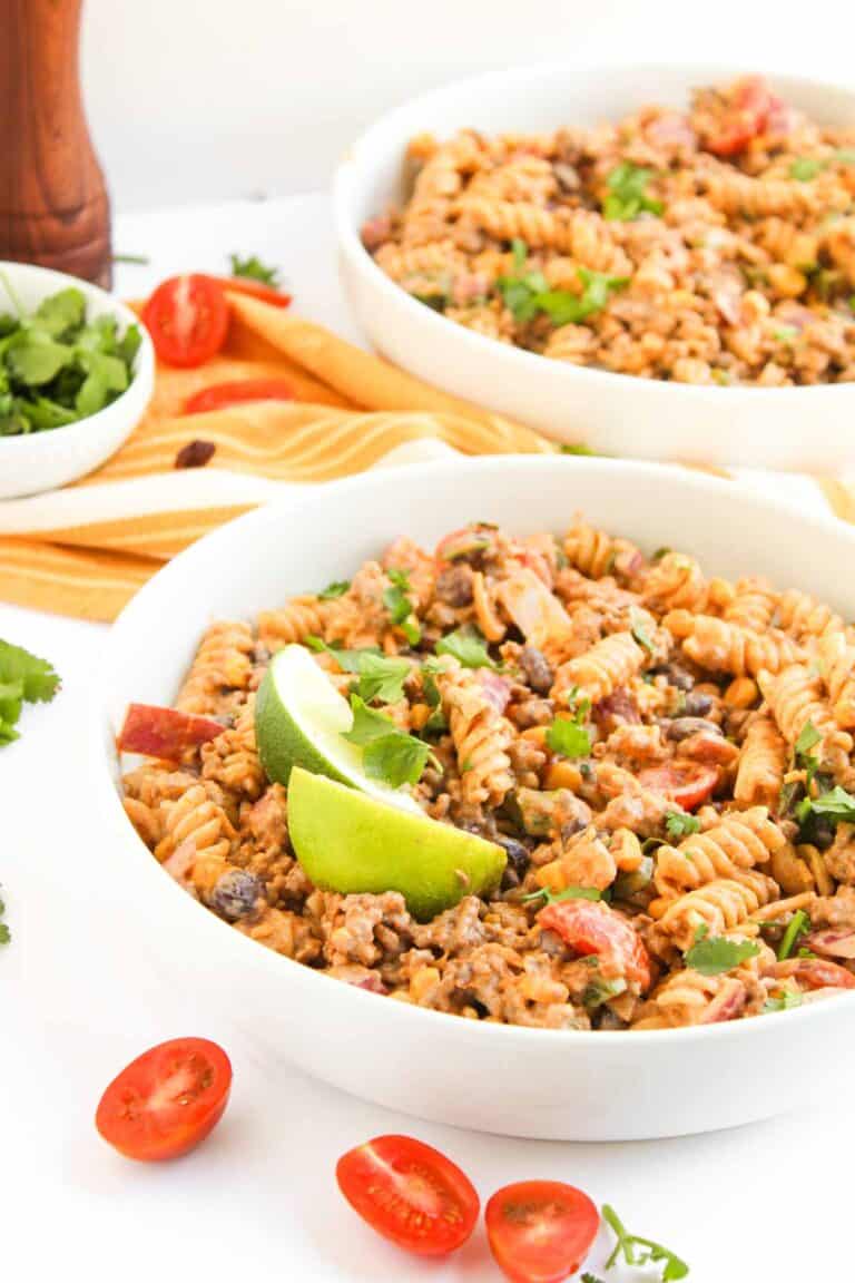 Two white bowls filled with creamy Taco Pasta Salad—rotini pasta mixed with ground meat, beans, tomatoes, and herbs, garnished with lime wedges, with sliced tomatoes and cilantro on the side.