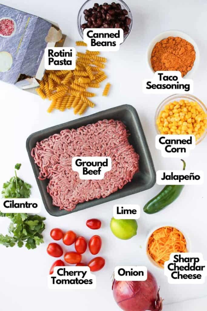 Top-down view of Taco Pasta Salad ingredients, featuring ground beef, rotini pasta, canned beans, taco seasoning, corn, jalape&ntilde;o, lime, cilantro, cherry tomatoes, onion, and cheddar cheese.
