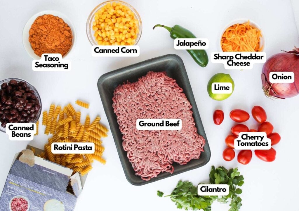 Ingredients for a Taco Pasta Salad dish, including ground beef, rotini pasta, canned beans, taco seasoning, corn, jalapeño, cheese, onion, lime, tomatoes, and cilantro, all arranged neatly on a white surface.
