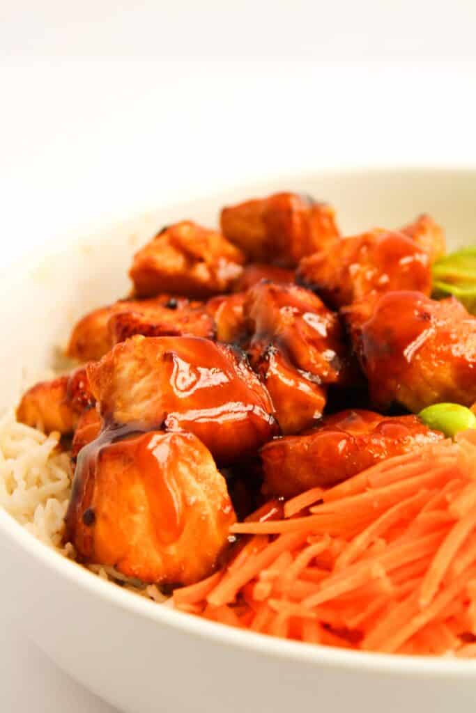 A bowl of rice crowned with savory teriyaki chicken pieces, complemented by sliced carrots and vibrant edamame beans.