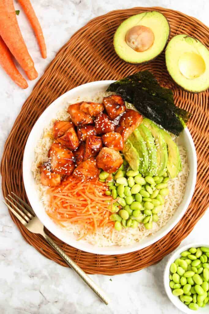 A bowl with rice, sliced avocado, glazed tofu, shredded carrots, edamame, and seaweed gives a nod to the classic Teriyaki Salmon Bowl. Halved avocado and whole carrots are placed nearby on a woven mat for an inviting presentation.