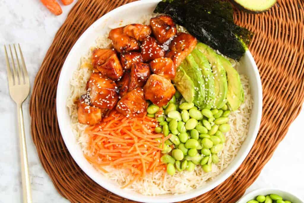A bowl filled with rice, teriyaki chicken, sliced avocado, edamame, shredded carrots, and nori is placed on a woven mat with a fork beside it. This enticing dish closely resembles a Teriyaki Salmon Bowl in its harmonious blend of flavors.