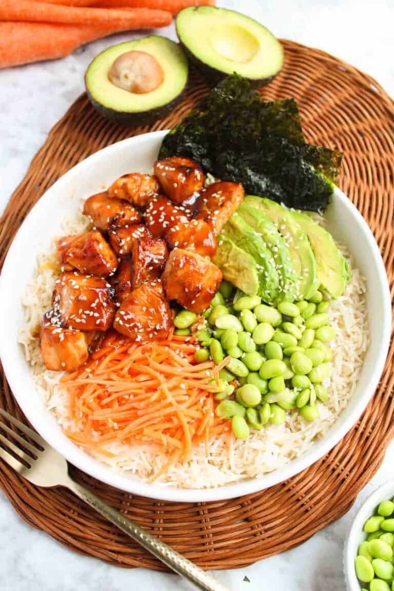A white bowl with rice, teriyaki glazed salmon, sliced avocado, shredded carrots, edamame, seaweed, and sesame seeds sits on a woven mat. In the background are halved avocados and carrots.