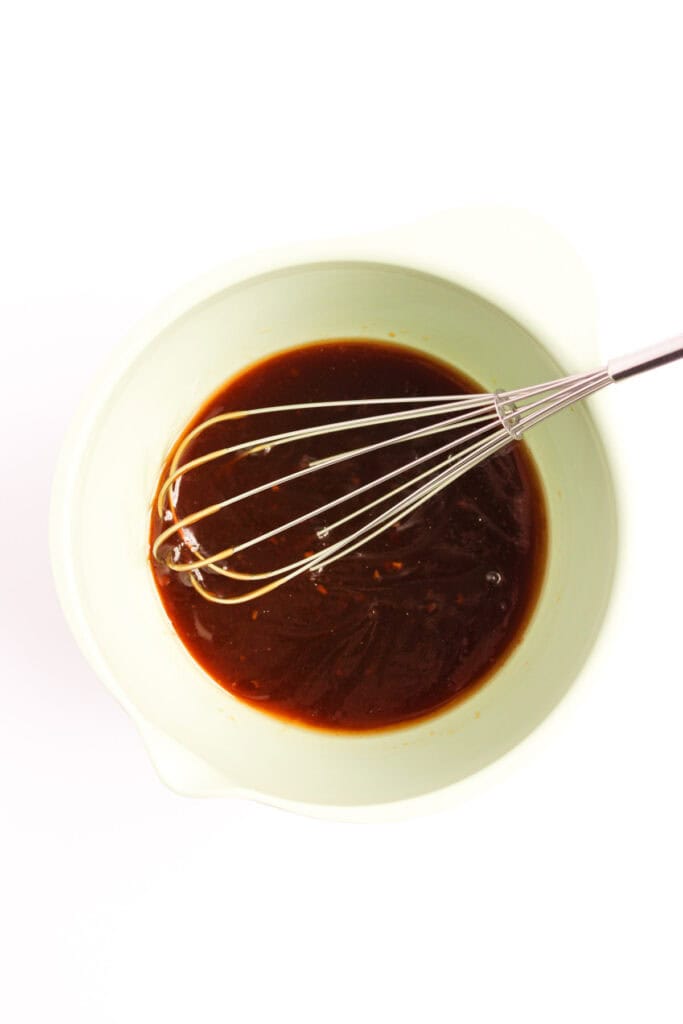 A whisk rests in a light-colored bowl of teriyaki sauce, perfect for marinating salmon, all set against a crisp white background.