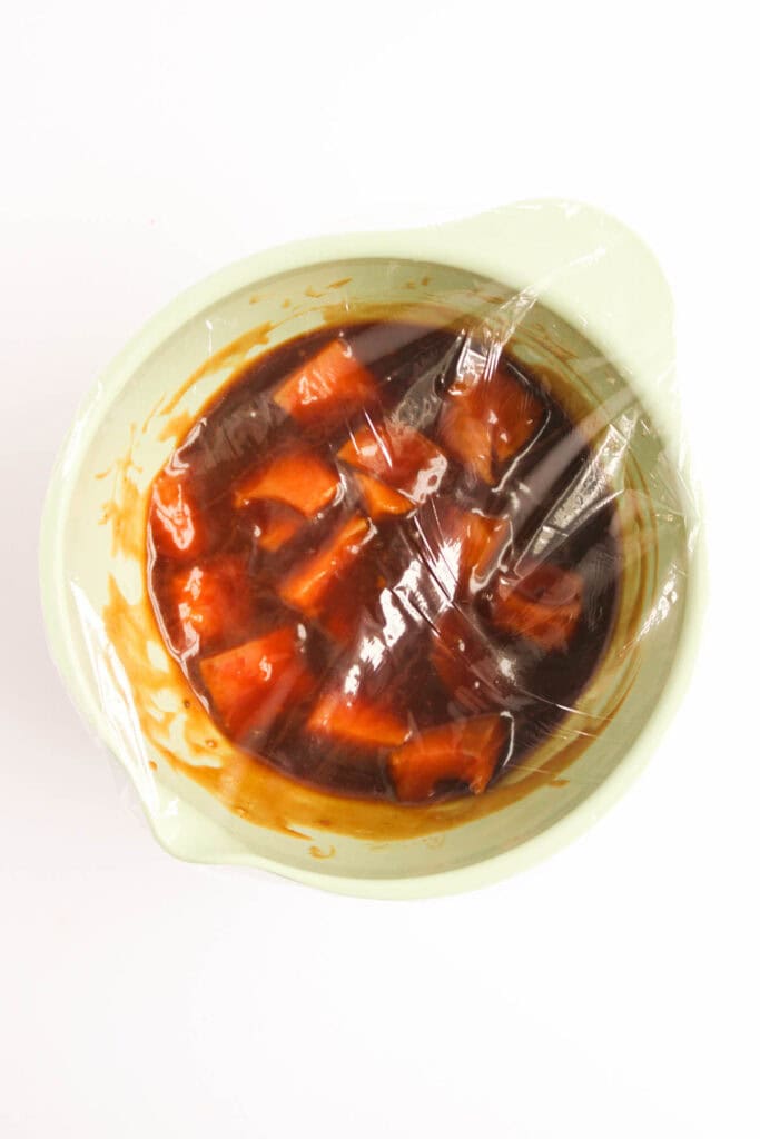 A bowl featuring diced orange pieces marinated in a teriyaki-inspired dark reddish sauce, covered with plastic wrap.