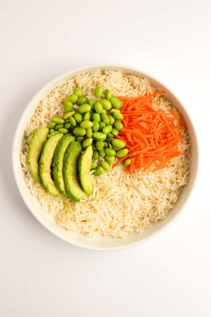 A bowl of white rice topped with sliced avocado, edamame, shredded carrots, and teriyaki salmon on a white background.