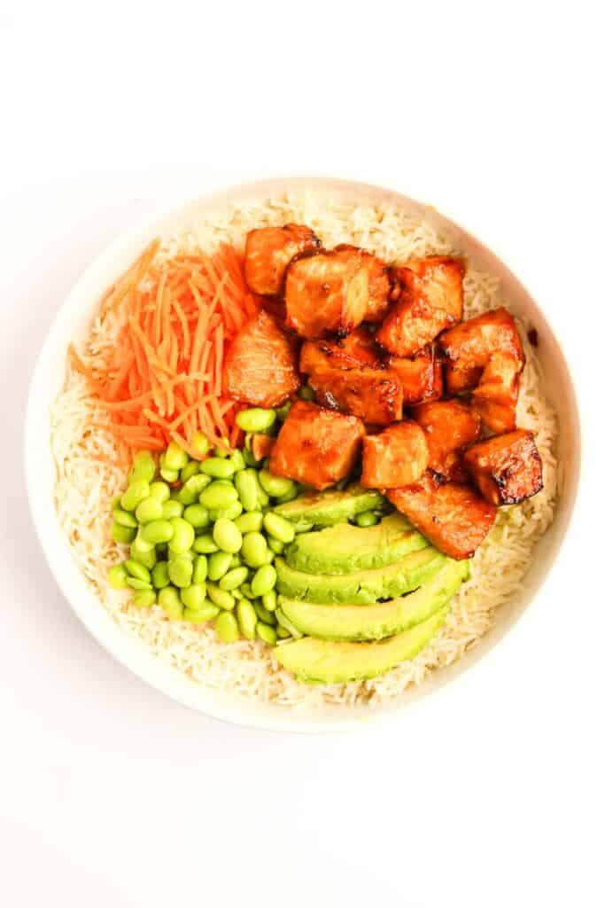 Teriyaki Salmon Bowl: A delightful rice bowl featuring succulent grilled chicken, sliced avocado, edamame, and shredded carrots, all arranged on a pristine white background.