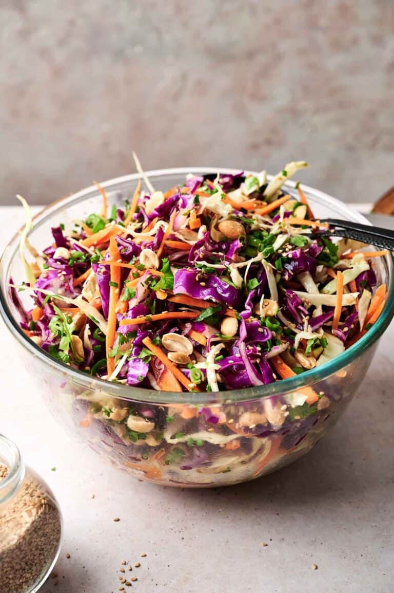 A glass bowl filled with vibrant Asian slaw, featuring shredded purple cabbage, carrots, green onions, and peanuts on a light surface.