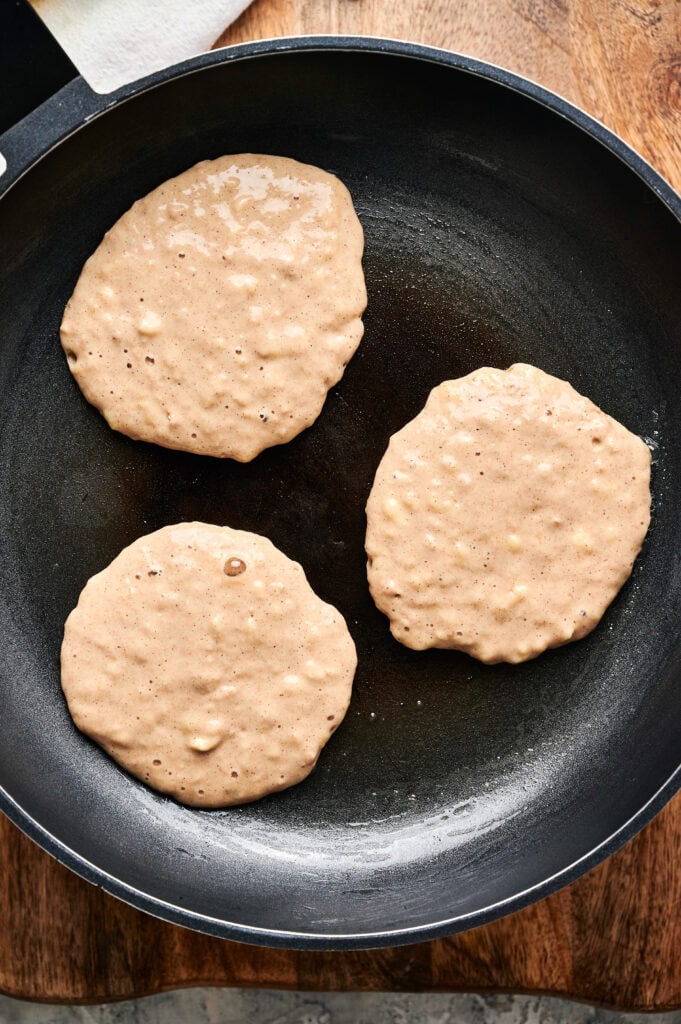 Three uncooked banana pancake batters are spread in a black frying pan, with bubbles forming on the surface, ready to be flipped.