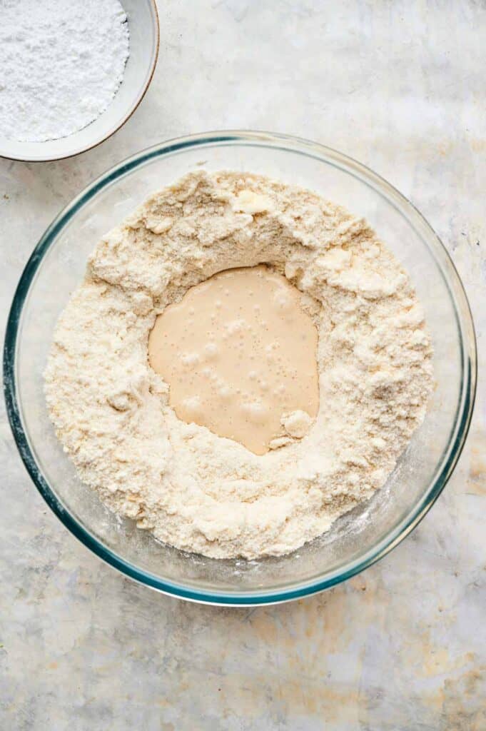 A glass bowl contains flour with a well in the center filled with a bubbling yeast mixture; nearby, another bowl of flour awaits, all set for baking delicious blueberry scones.