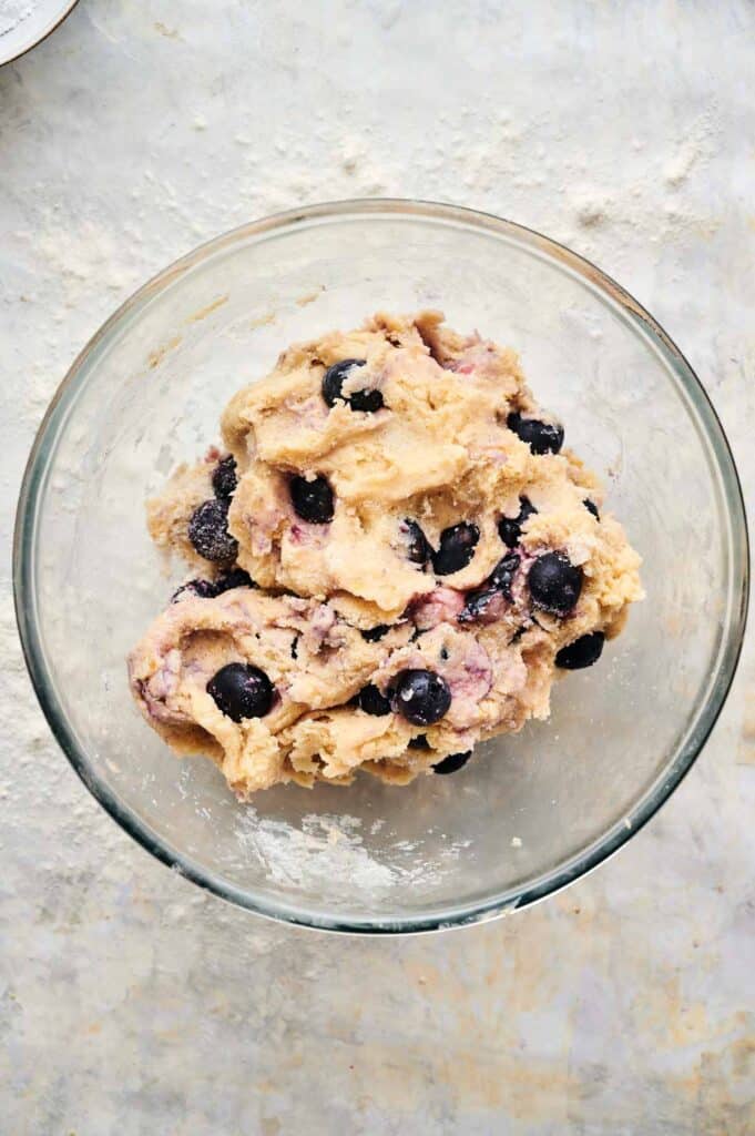 A glass bowl containing raw dough for blueberry scones, mixed with fresh blueberries, sits on a light-colored surface.