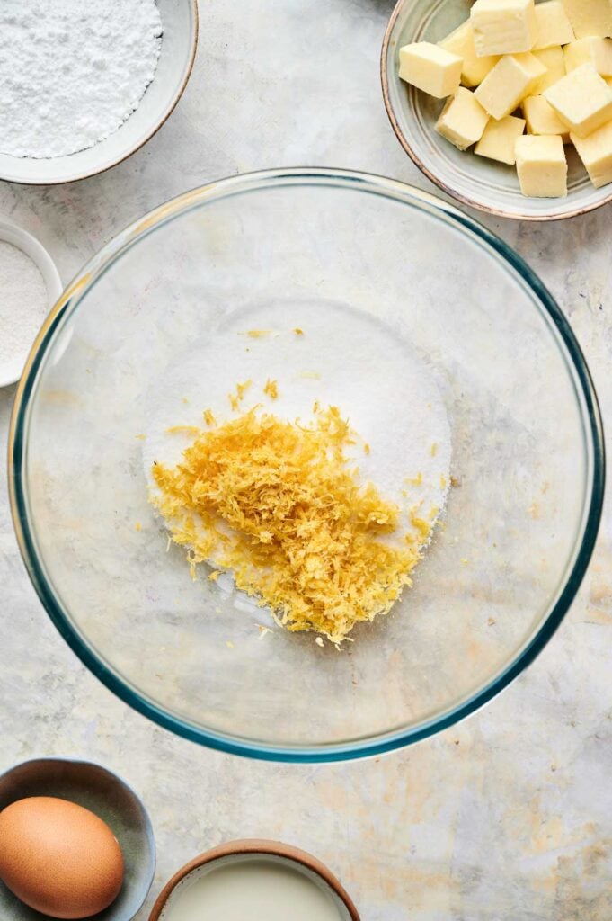 A glass mixing bowl with sugar and lemon zest, surrounded by bowls of cubed butter, powdered sugar, flour, an egg, and milk—perfect ingredients for baking delicious blueberry scones.
