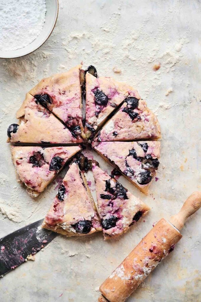 Round blueberry scone cut into eight wedges on a floured surface, with a rolling pin and knife beside it&mdash;perfect for making homemade blueberry scones.