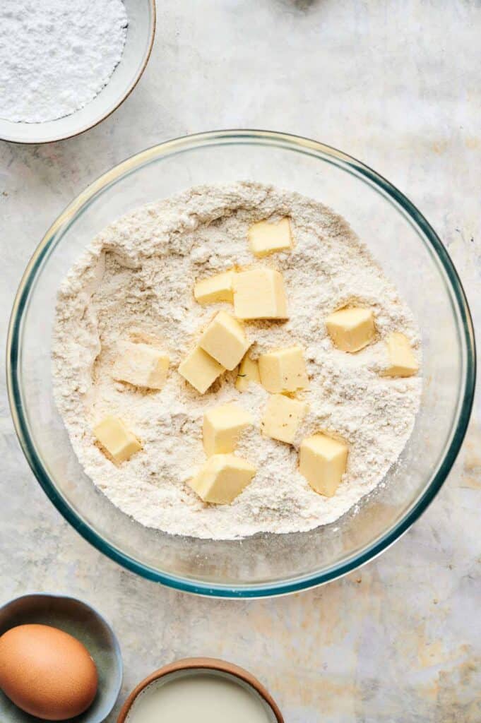 A glass bowl with flour and cubed butter, surrounded by a bowl of powdered sugar, an egg, milk, and ingredients ready to create delicious blueberry scones.