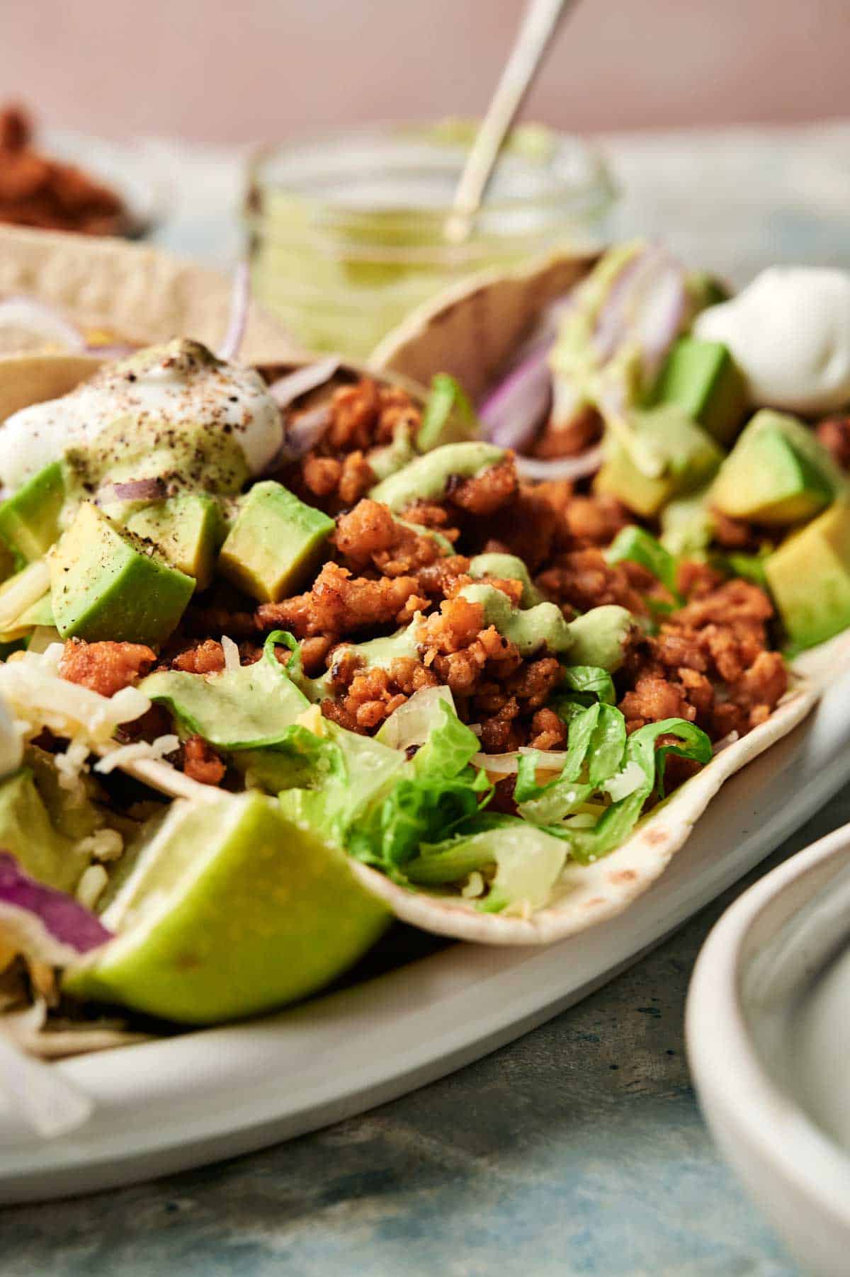 Close-up of a chicken taco filled with seasoned meat, diced avocado, lettuce, onions, sour cream, and a lime wedge on the side.