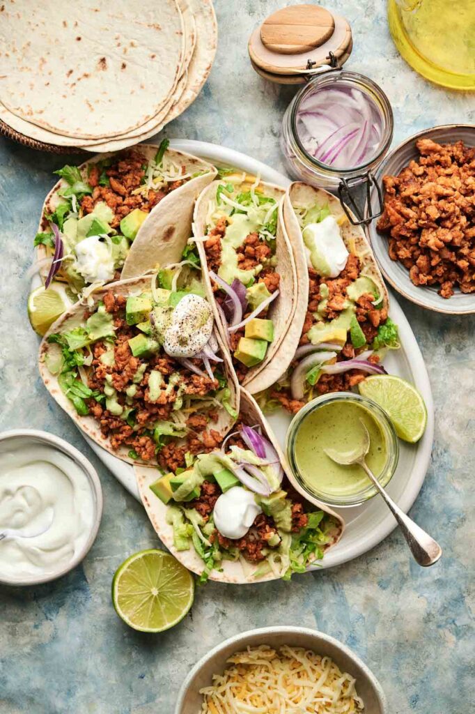 Chicken Tacos with seasoned crumbled filling, lettuce, avocado, red onion, sour cream, and lime wedges, surrounded by bowls of toppings and tortillas on a light blue surface.
