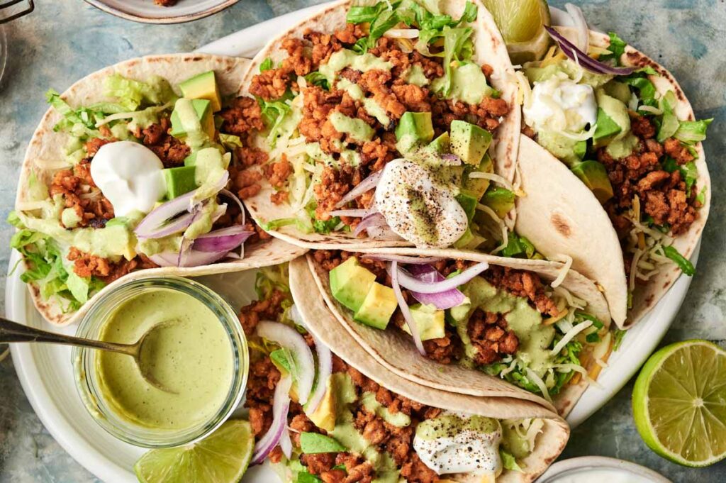 Chicken Tacos filled with ground meat, avocado, lettuce, red onion, shredded cheese, and sauce are arranged on a plate with lime wedges and a jar of green sauce nearby.