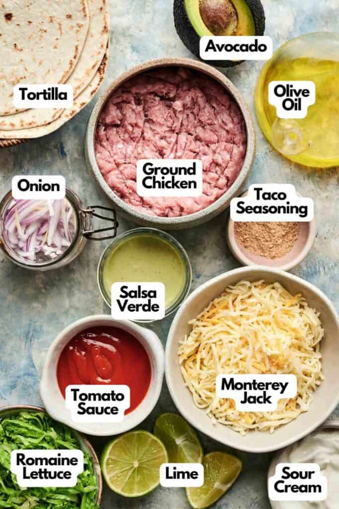Ingredients for chicken taco night arranged on a table, including tortilla, ground chicken, taco seasoning, cheese, salsa verde, tomato sauce, onion, olive oil, avocado, lime, lettuce, and sour cream.