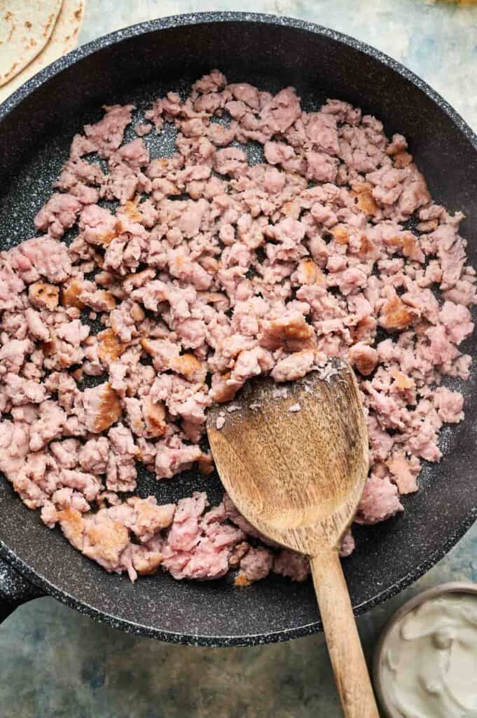 Ground chicken taco meat is being cooked in a skillet, with a wooden spoon stirring the meat.