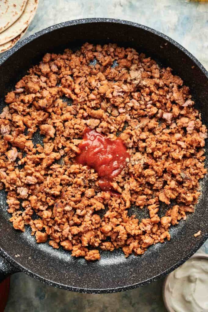 Cooked ground chicken taco meat with seasoning and a dollop of red sauce in a black skillet, viewed from above.