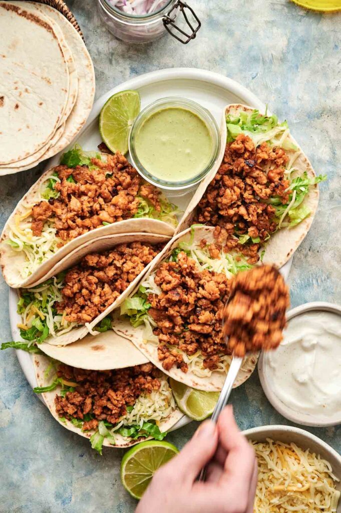 A hand spoons savory chicken taco filling onto a plate of chicken tacos with tortillas, lettuce, cheese, lime wedges, and two sauces visible.