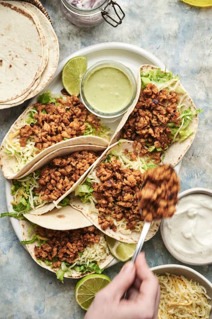 A plate of soft chicken tacos filled with seasoned meat, lettuce, and cheese, served with lime wedges and dipping sauces. A hand is spooning savory chicken onto one taco.