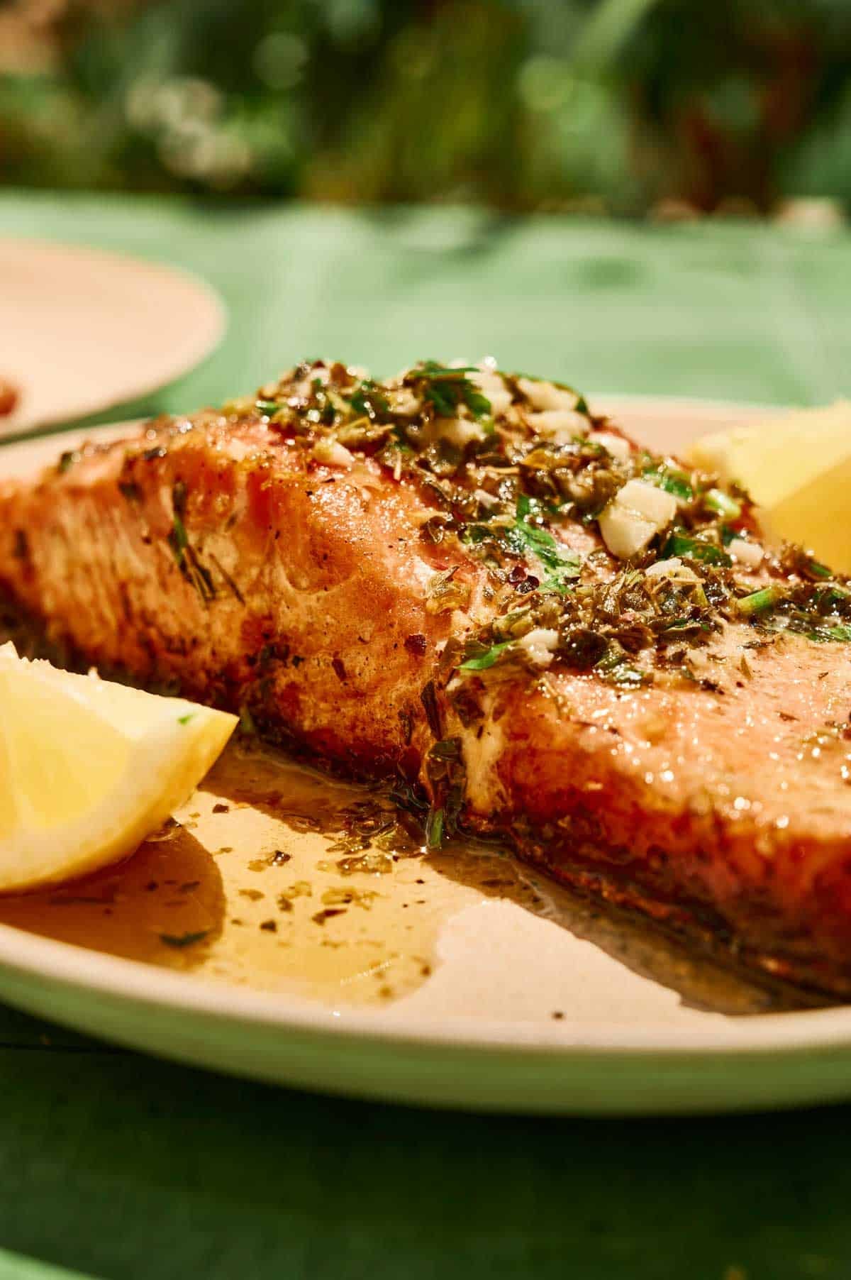 A close-up of a grilled salmon fillet topped with herbs and sauce, served on a plate with lemon wedges.