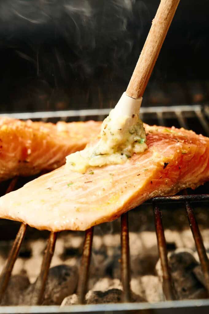 A close-up of grilled salmon fillets on a grill being brushed with a herb butter mixture using a silicone brush.