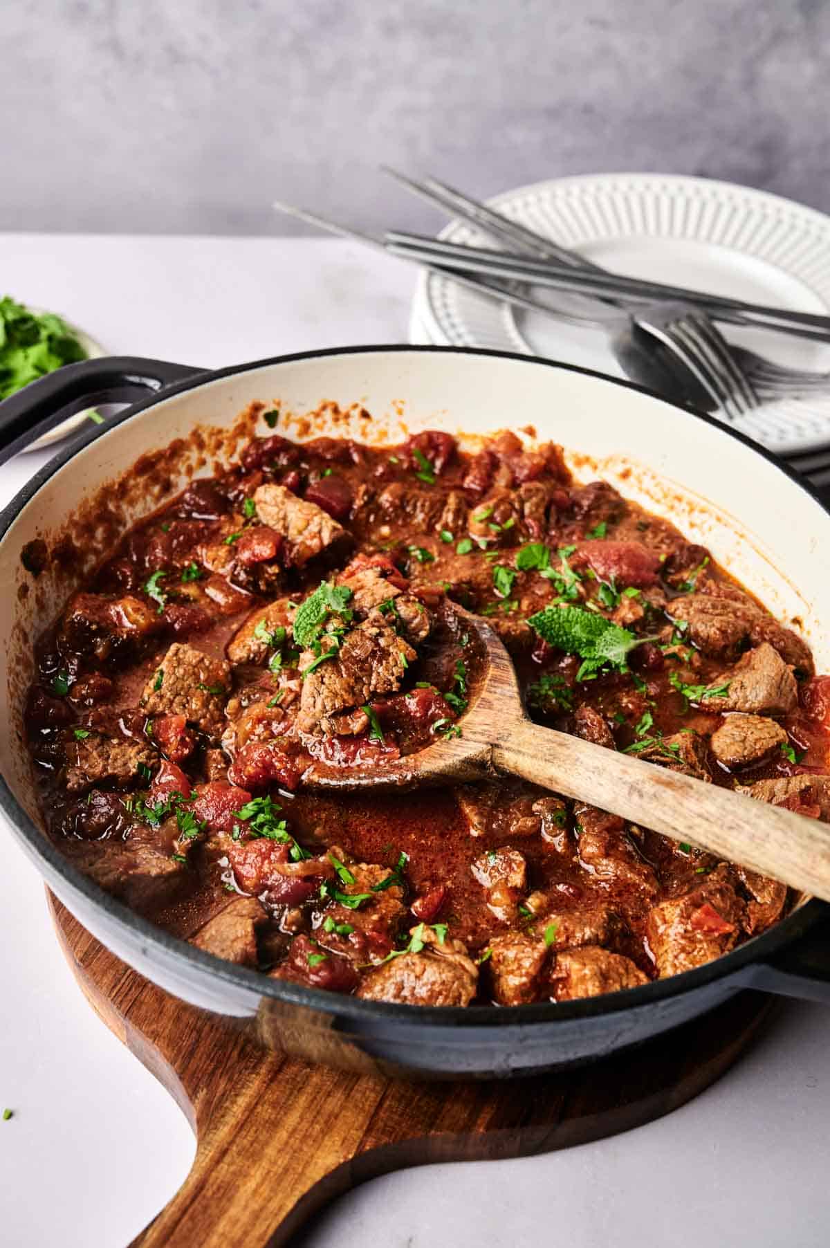 A pot of hearty beef and lamb stew with tomato sauce and herbs, garnished with chopped parsley, with a wooden spoon, plates, and cutlery in the background.