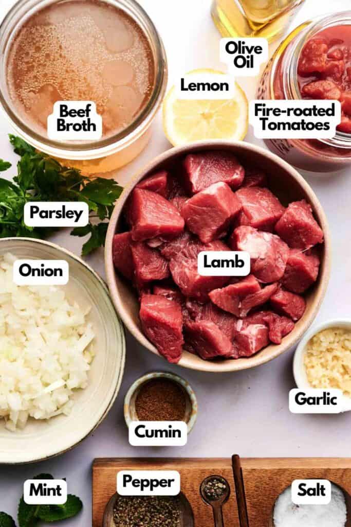 Top-down view of lamb stew ingredients: cubed lamb, chopped onion, beef broth, fire-roasted tomatoes, olive oil, lemon, parsley, garlic, cumin, mint, salt, and pepper&mdash;all you need for a delicious homemade lamb stew.