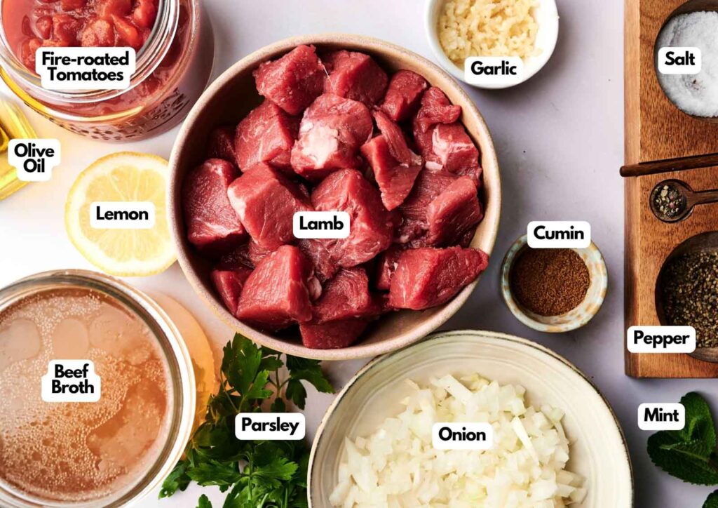 Labeled ingredients for a lamb stew recipe are arranged on a table, including cubed lamb, fire-roasted tomatoes, garlic, cumin, salt, pepper, onion, parsley, beef broth, lemon, olive oil, and mint.