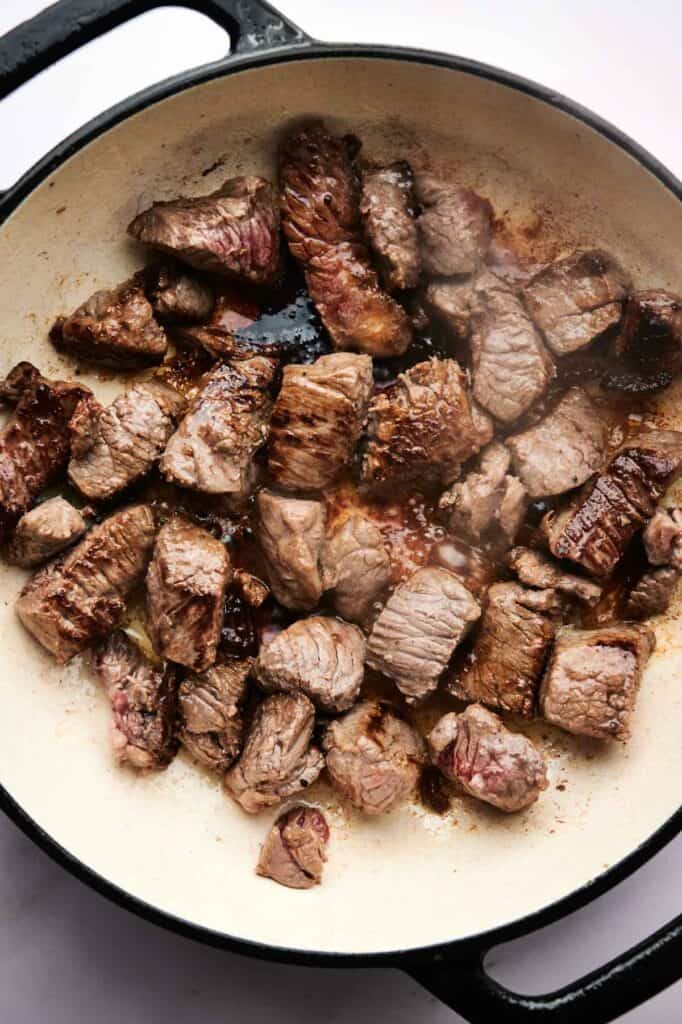Chunks of beef, perfect for a hearty lamb stew, are browning in a white pot, with some pieces showing seared edges and others still partially raw.