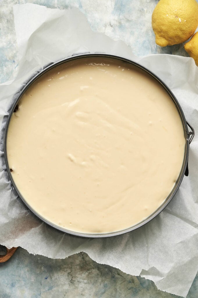 A round baking pan lined with parchment paper is filled with uncooked lemon cheesecake batter. Lemons are visible in the top right corner.