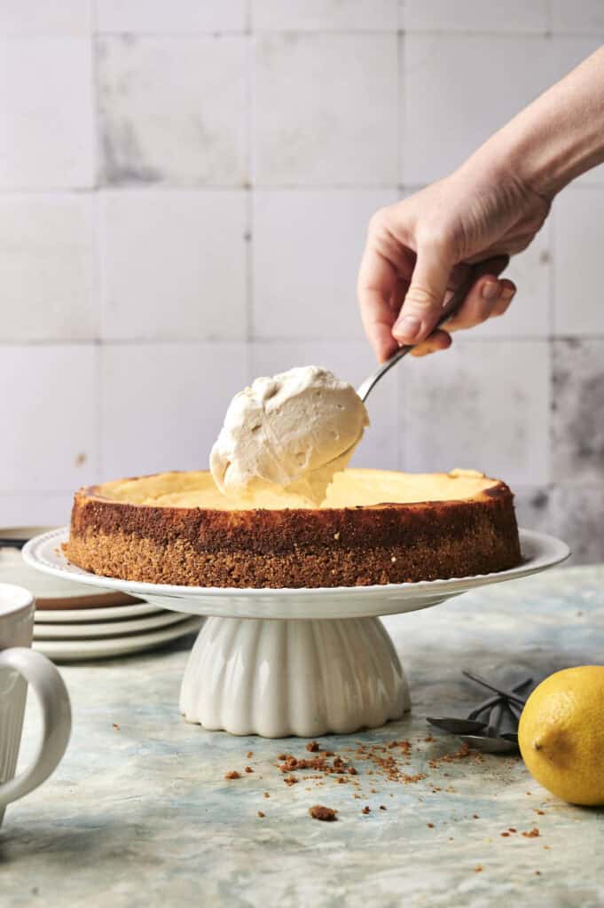 A hand places a scoop of whipped cream on top of a lemon cheesecake displayed on a white cake stand. Plates, a cup, and a fresh lemon are arranged nearby.