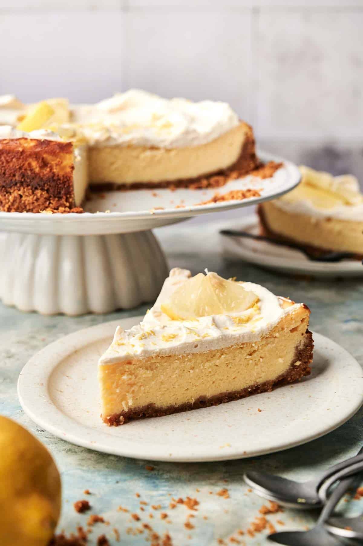 A slice of lemon cheesecake topped with whipped cream and a lemon slice sits on a plate, with the remaining cake on a stand in the background.