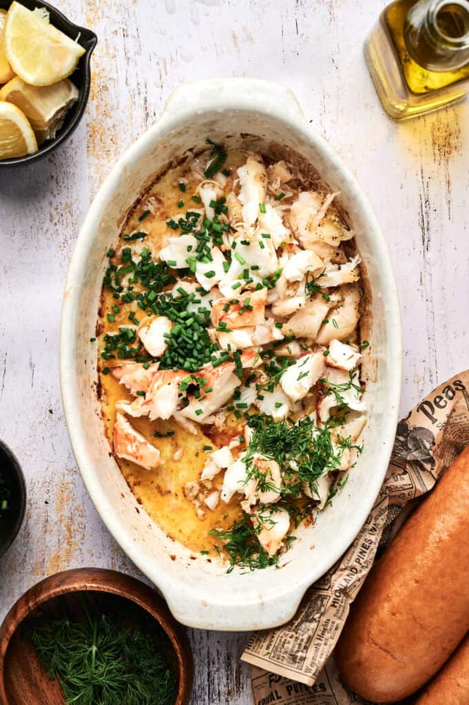 Oval baking dish filled with baked fish topped with chopped herbs, surrounded by lemon wedges, a bread loaf, olive oil, a lobster roll, and a bowl of fresh greens on a white surface.