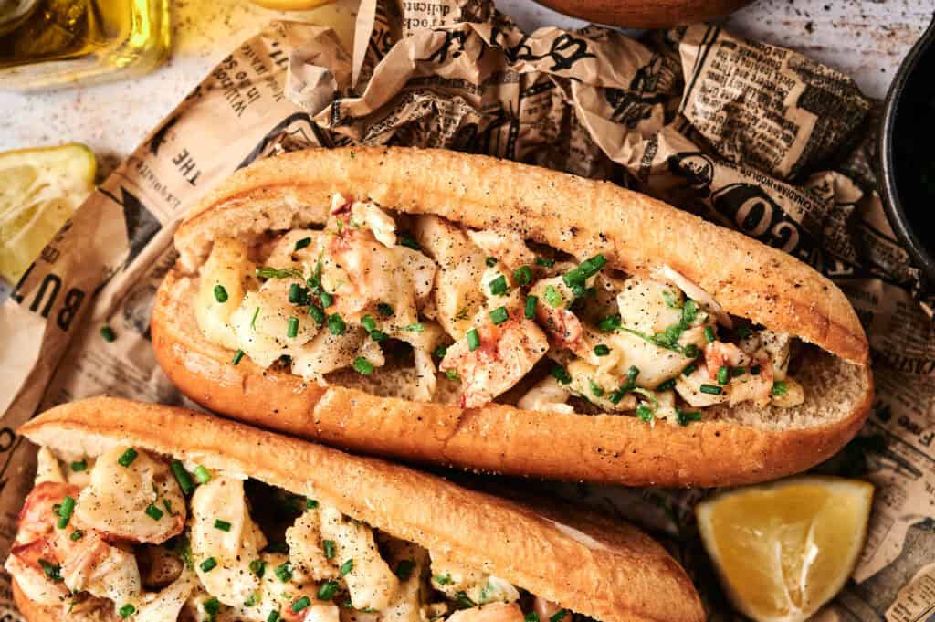 Two lobster rolls topped with chopped chives sit on crumpled newspaper, with lemon wedges nearby—perfect for any lobster roll lover seeking a classic seaside treat.
