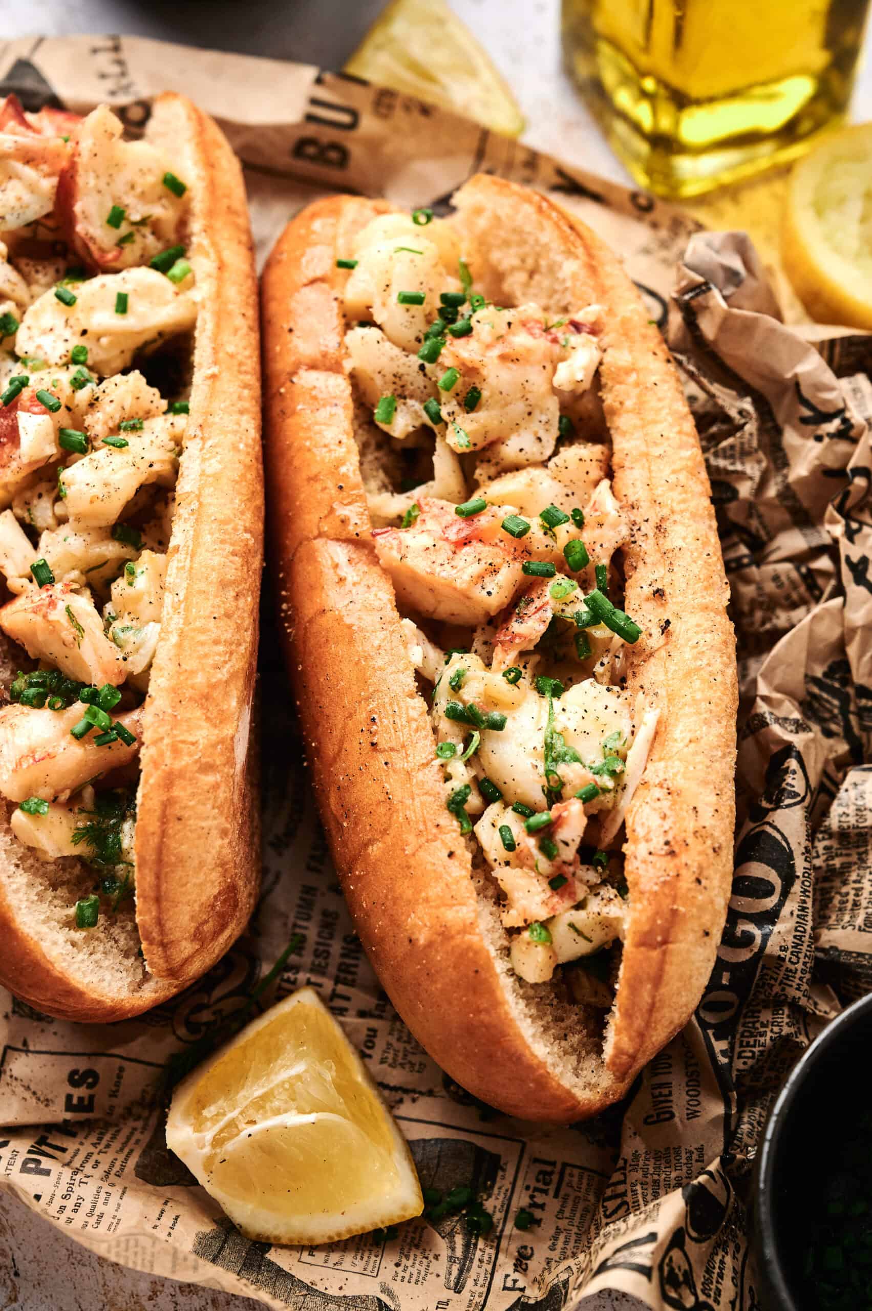 Two classic lobster rolls on toasted buns, topped with chives and served on newspaper-style paper, bring the iconic lobster roll experience to life. Lemon wedges are included on the side for a fresh twist.
